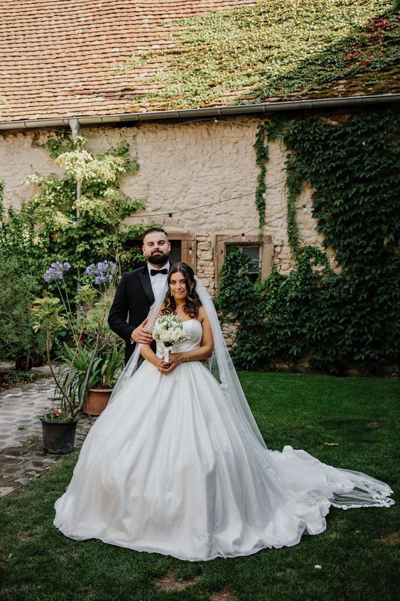 A bride and groom pose together on the lawn of a rustic venue with ivy-covered stone walls in the background.