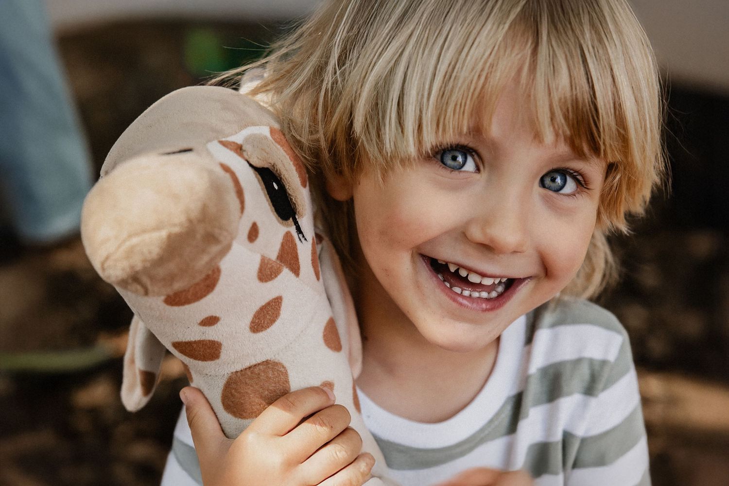 Smiling blonde child with blue eyes hugging a giraffe stuffed animal toy, wearing a striped shirt.