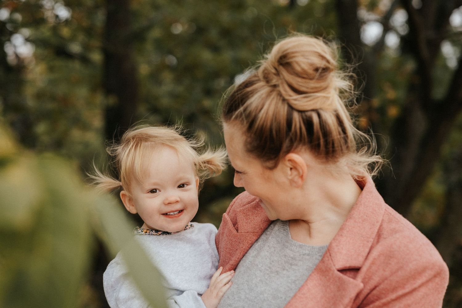 A mother tenderly holds her smiling toddler outdoors, both with blonde hair, surrounded by autumn foliage.