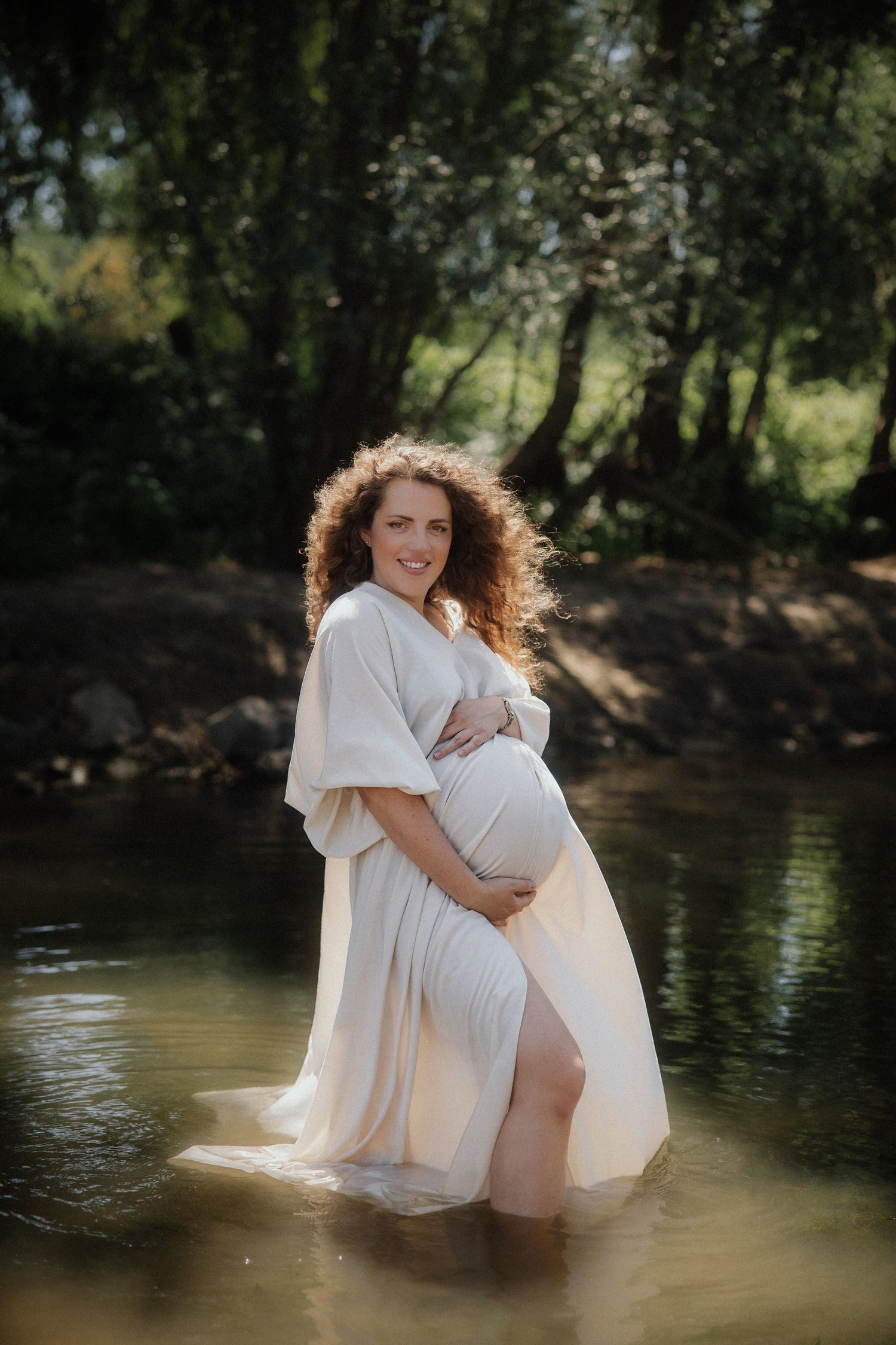Woman in white dress sits peacefully by a serene stream in a natural outdoor setting.