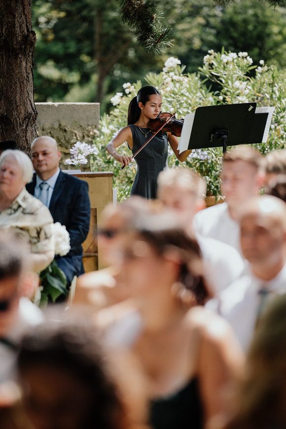 A violinist performs during an outdoor wedding ceremony with guests seated and floral decorations in the background.
