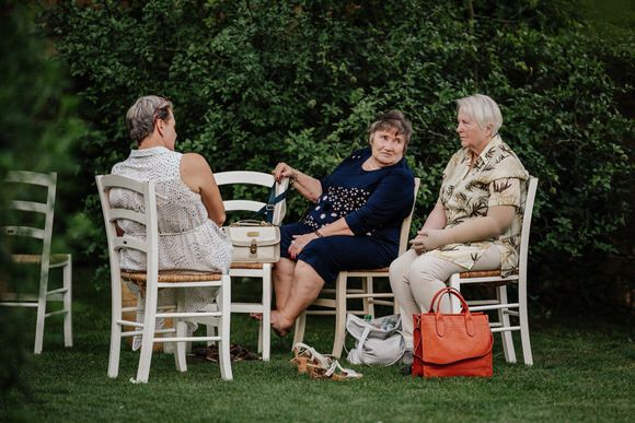 Hochzeitsfotograf Boudier & Koeller – Drei Personen unterhalten sich auf weißen Holzstühlen im Garten.