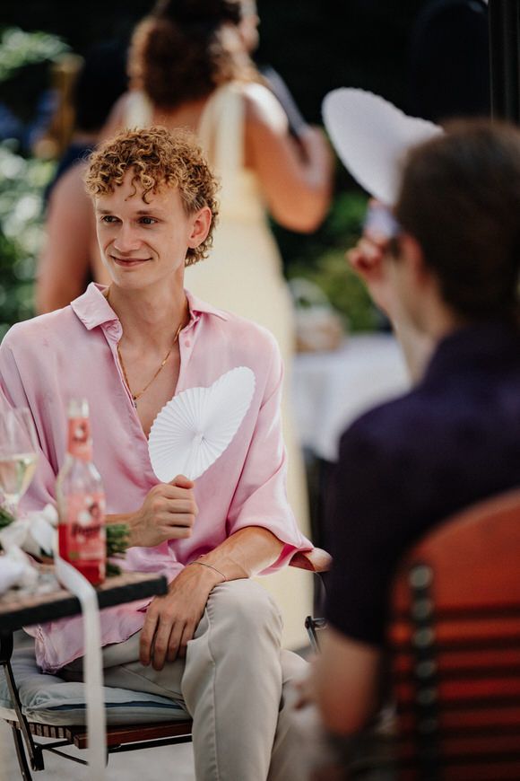 A person in a pink button-down shirt and khaki pants sits at an outdoor restaurant table smiling during a social gathering.