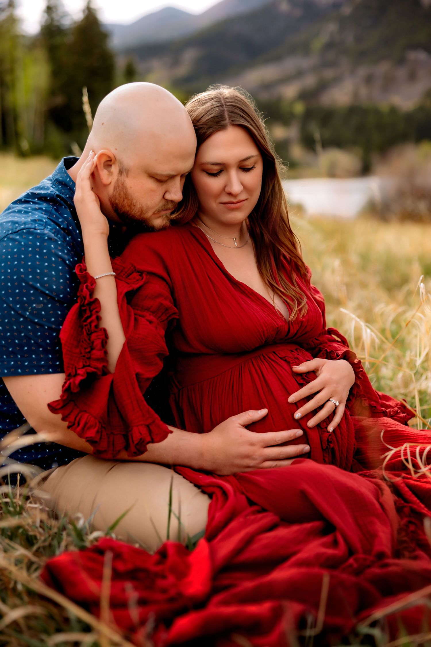 Couple sharing tender maternity moment outdoors, woman in red dress cradling baby bump, mountains in background.