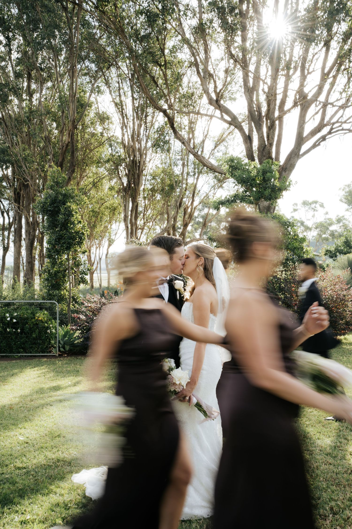Bride and groom share a kiss outdoors while blurred bridesmaids in dark gowns walk past during a garden wedding ceremony.
