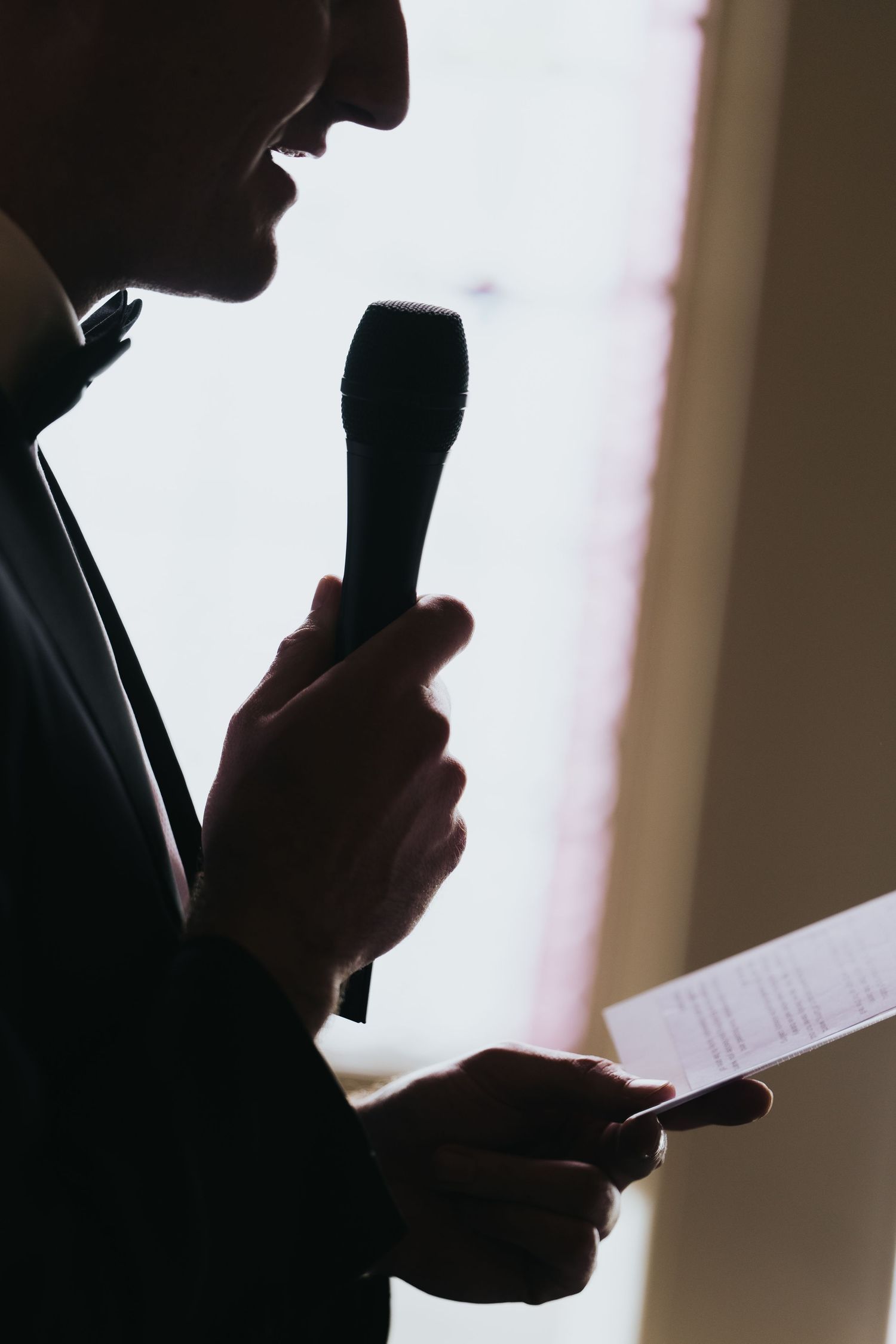 Silhouette of a man holding a microphone and paper, speaking at an event in dramatic backlit lighting.