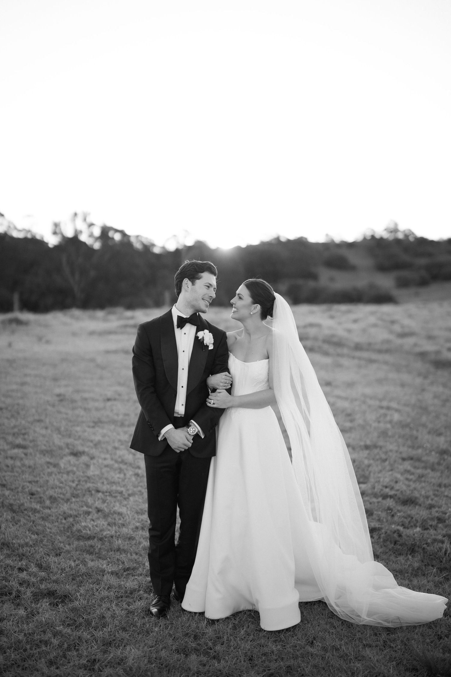 Black and white portrait of bride and groom embracing in an open field at sunset on wedding day.