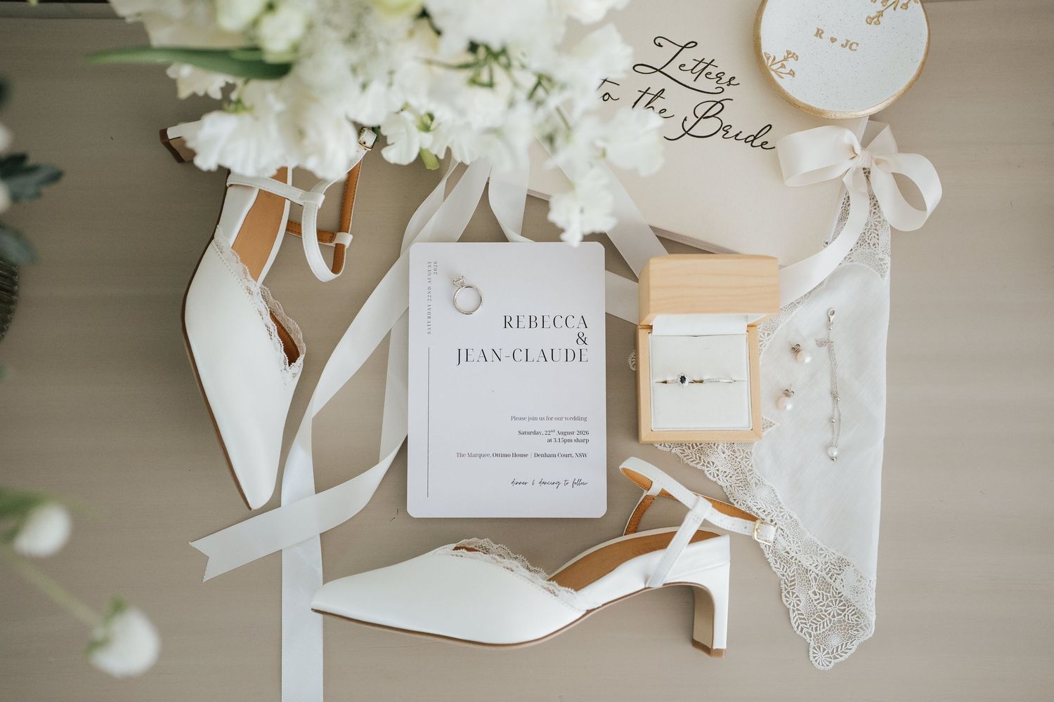 Elegant wedding flat lay featuring white heels, invitation for Rebecca and Jean-Claude, ring box, and floral accents.