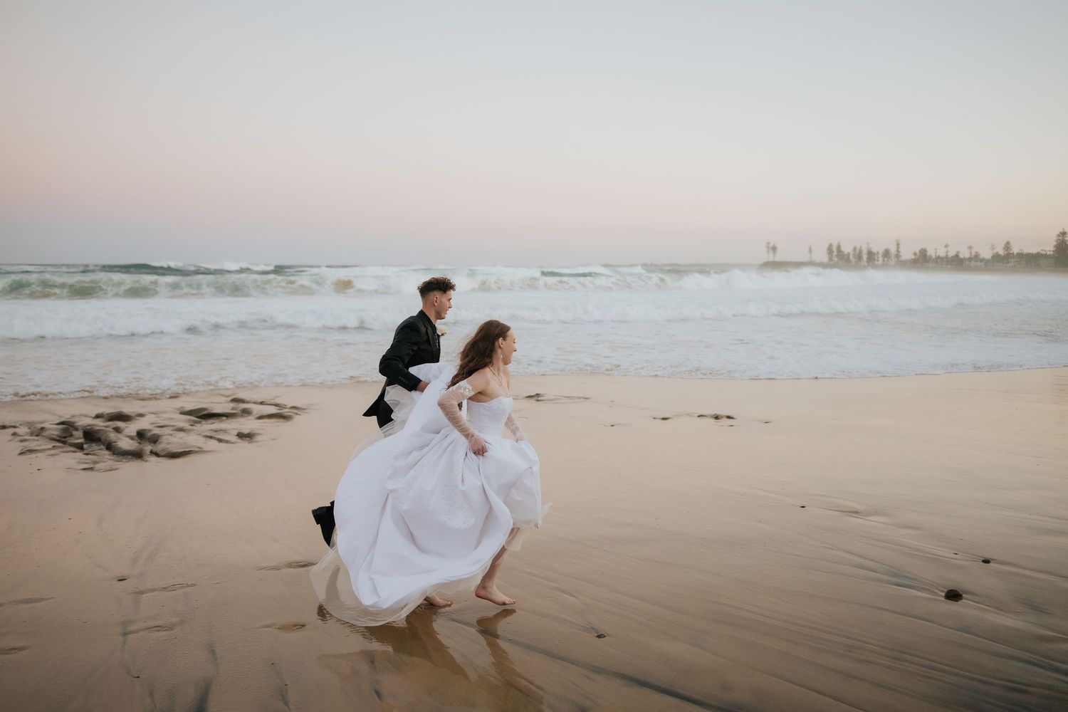 Bride and groom running on a sandy beach at sunset with ocean waves in the background.