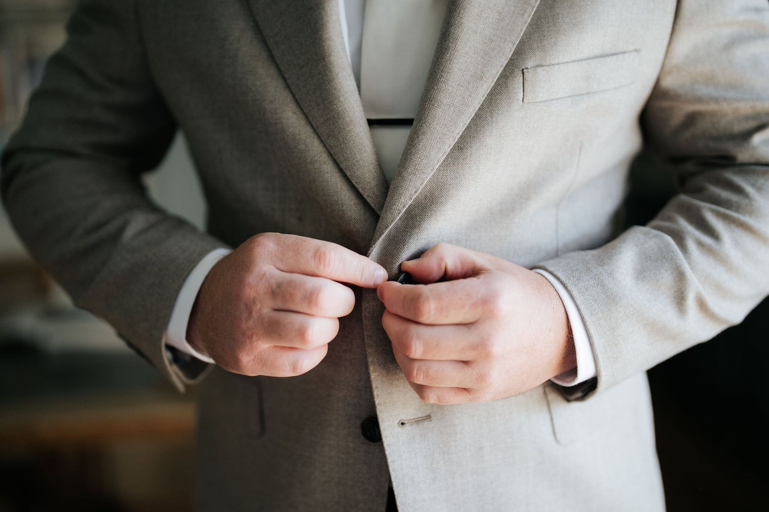 Groom buttoning his gray wedding suit jacket while getting dressed on his wedding day.