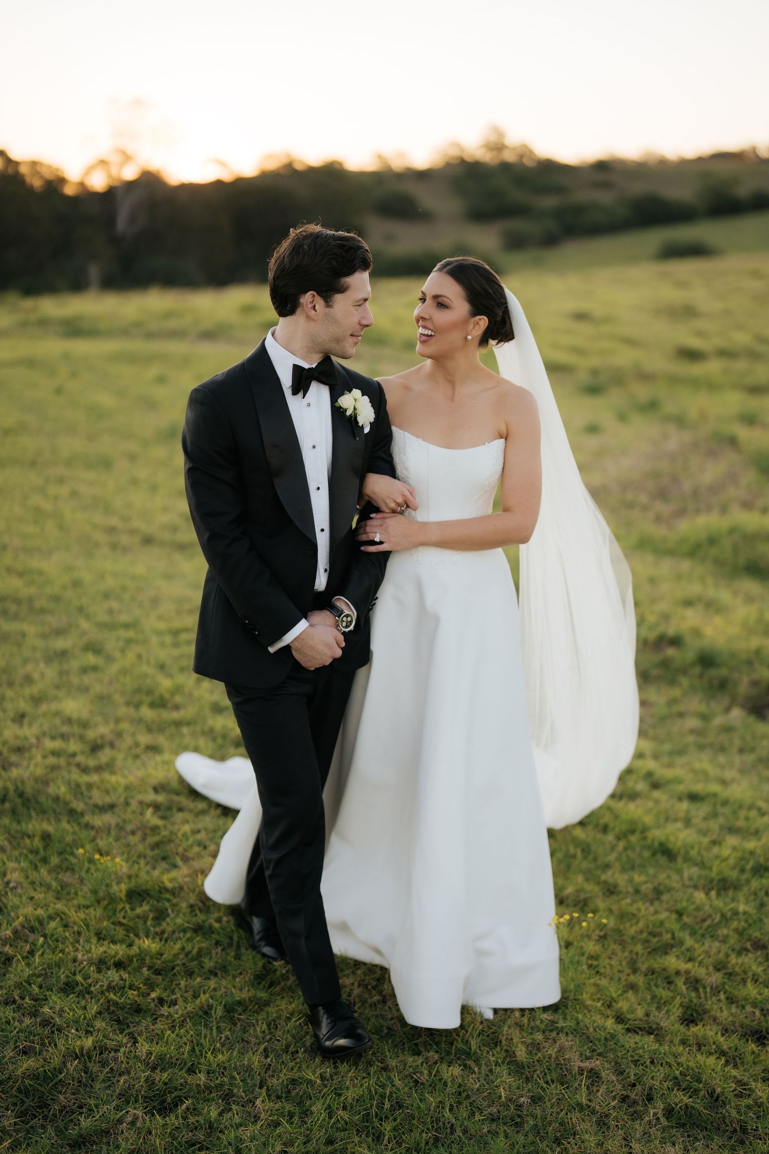 Bride and groom walking together through green field at sunset, sharing a joyful moment on wedding day.