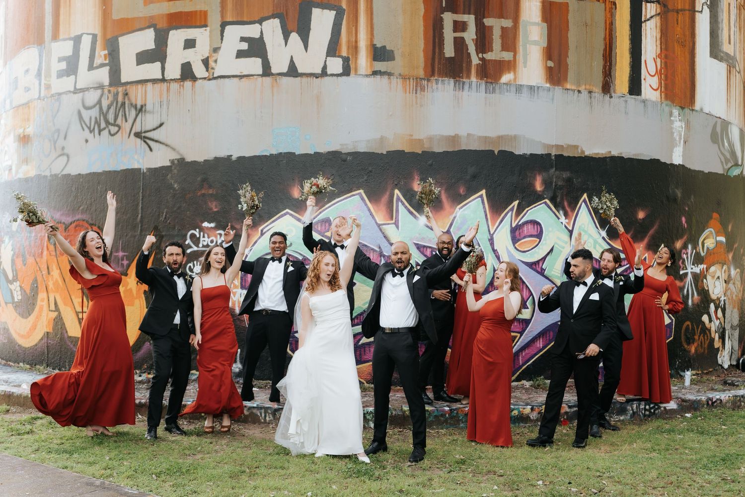 Wedding party in red dresses and tuxedos celebrating in front of a graffiti wall.