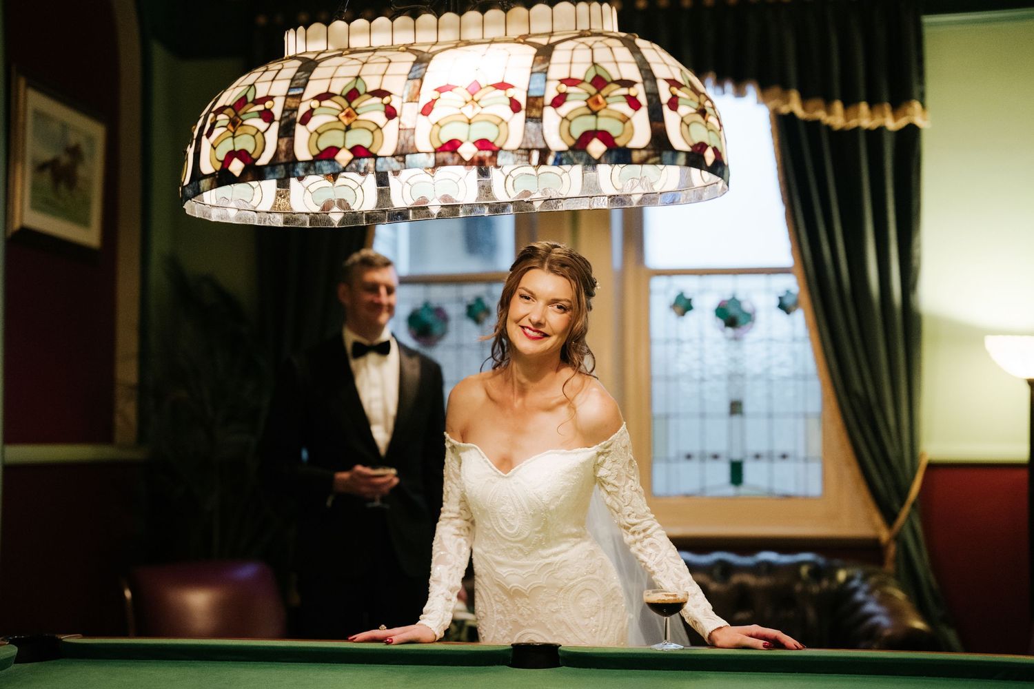 Bride smiling at billiard table in elegant wedding dress with groom in tuxedo standing behind her near stained glass window.