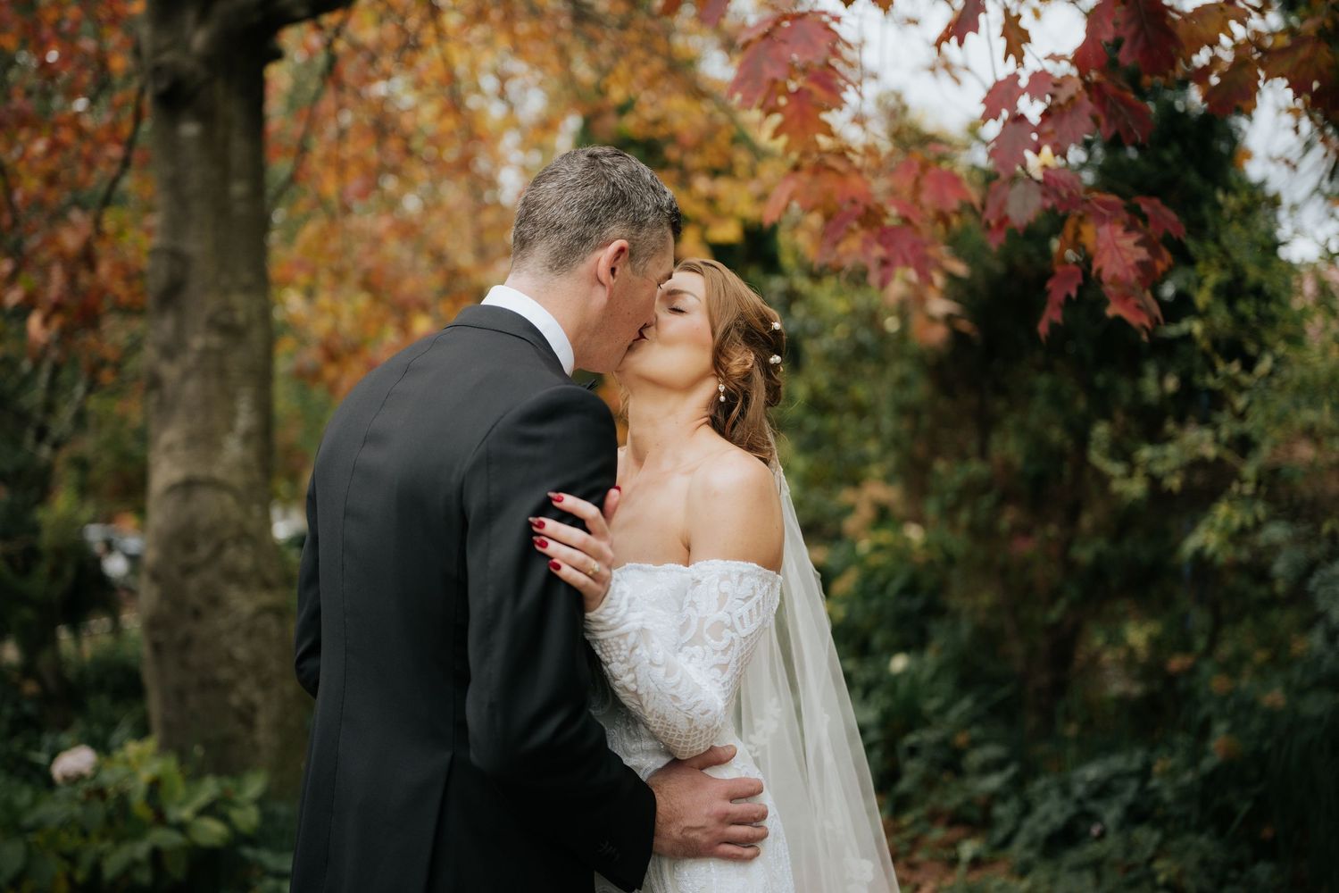 Bride and groom sharing a romantic kiss outdoors surrounded by colorful autumn foliage in a garden setting.