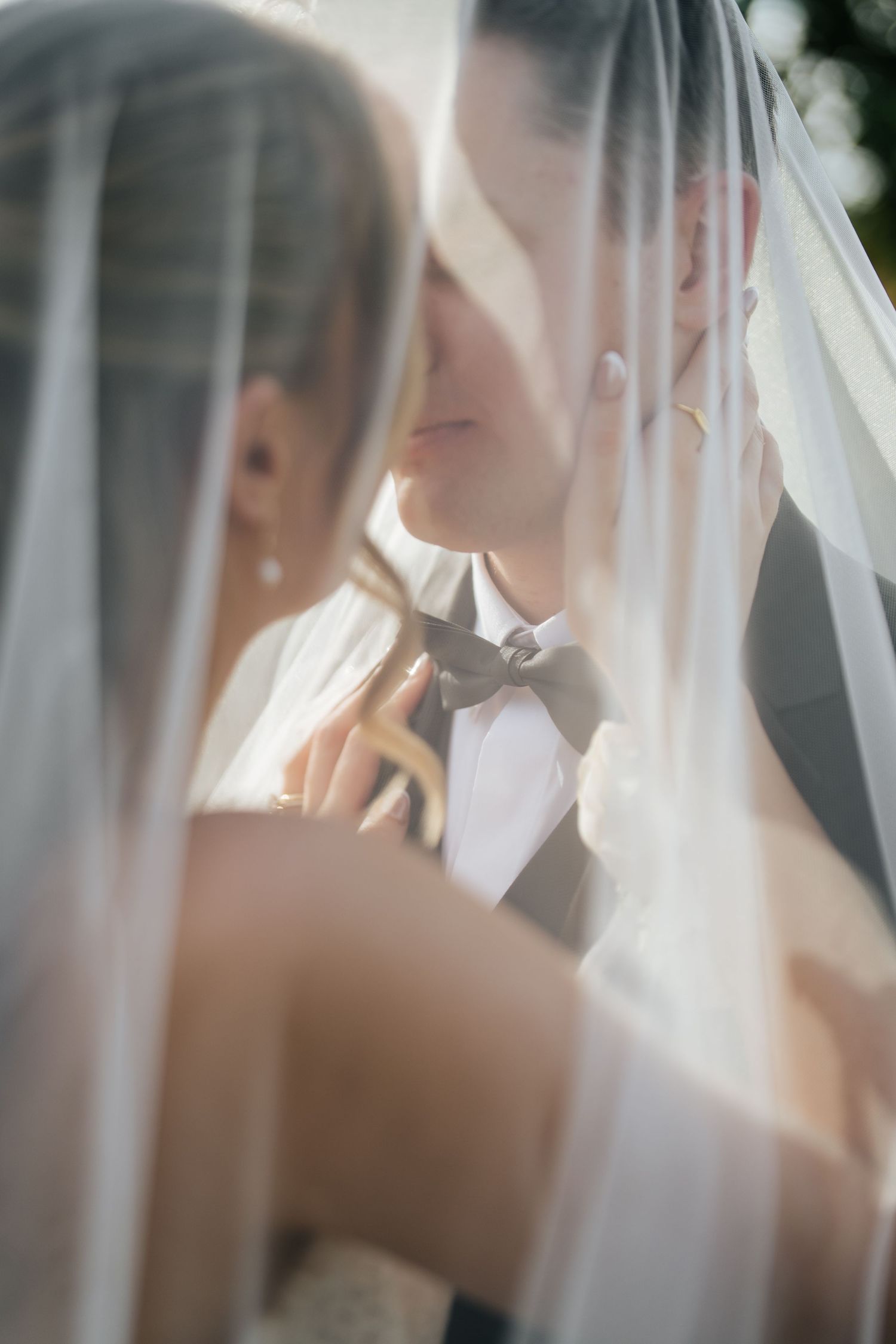 Bride and groom sharing a tender kiss under a flowing white wedding veil in soft natural light.