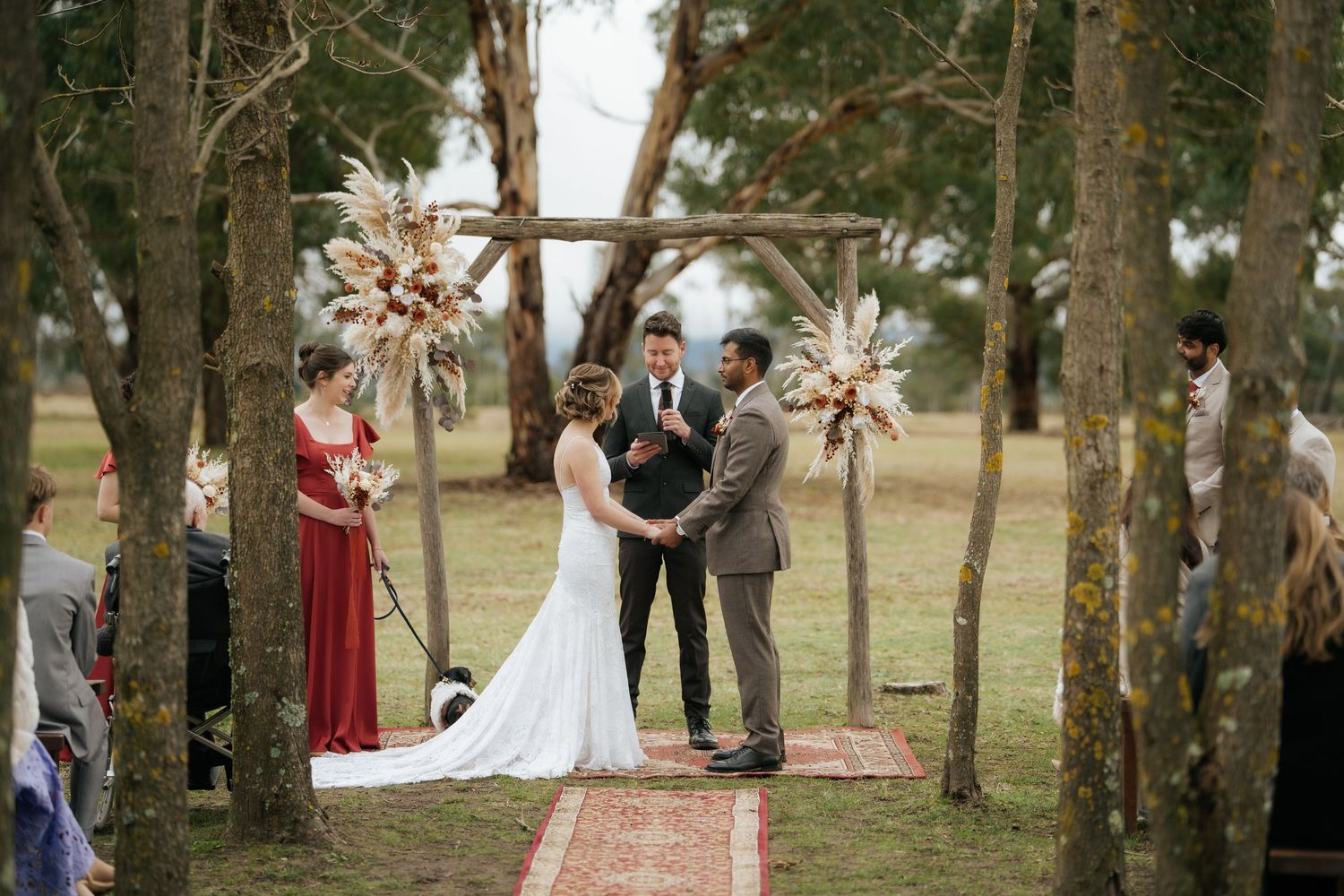 Outdoor wedding ceremony under rustic wooden arch with pampas grass, bride and groom holding hands with officiant in wooded setting.