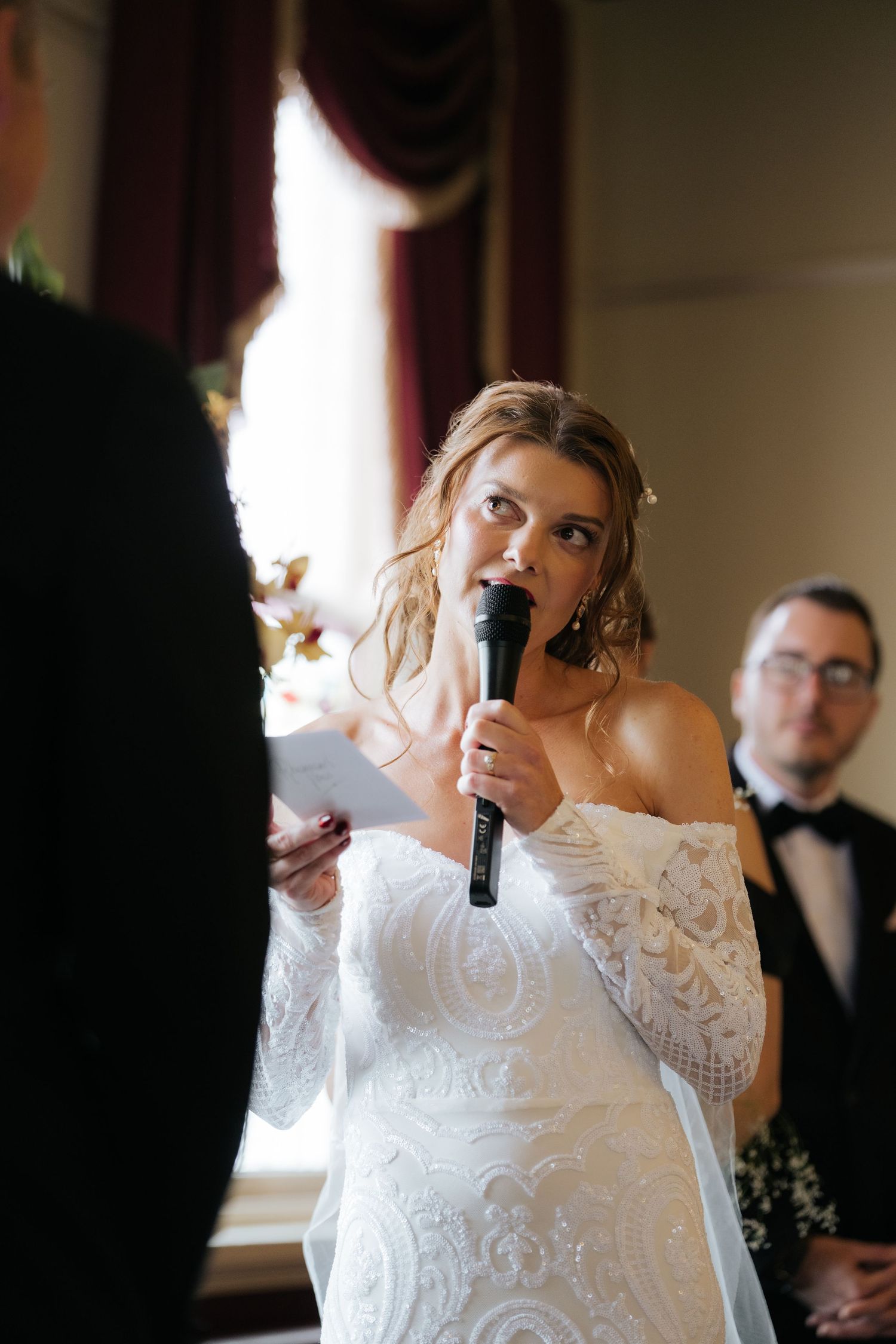 Bride in lace off-shoulder gown reads wedding vows into microphone during elegant indoor ceremony.