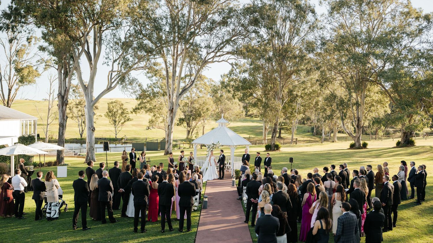 Outdoor wedding ceremony on lush green lawn surrounded by tall eucalyptus trees with guests gathered around.