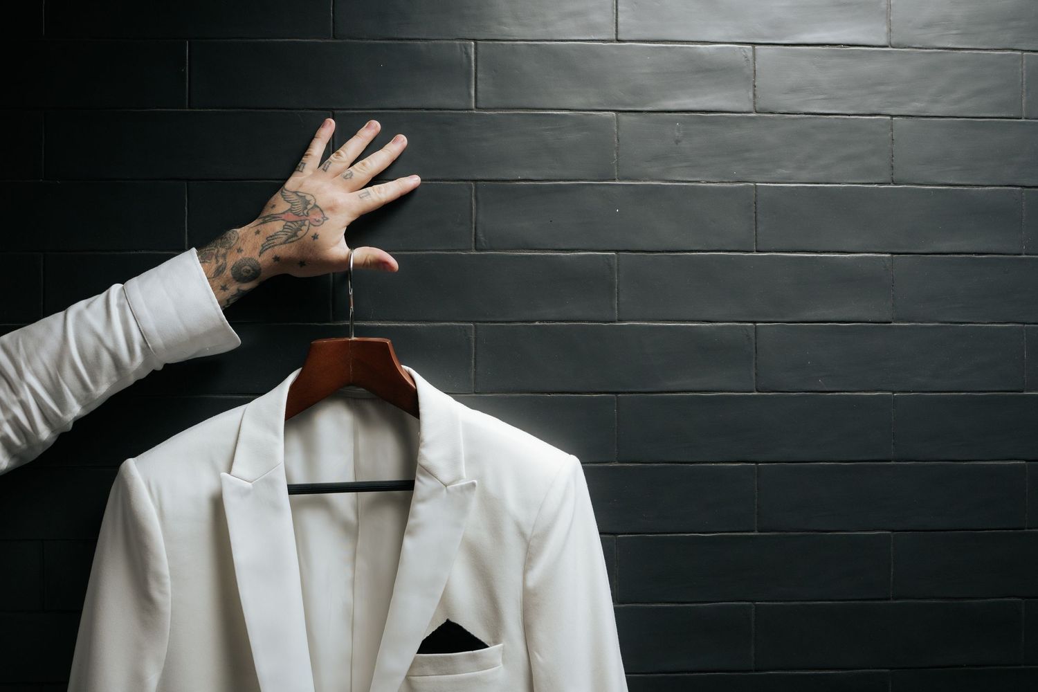 Tattooed hand holding a white wedding suit jacket on a wooden hanger against a dark tile wall.