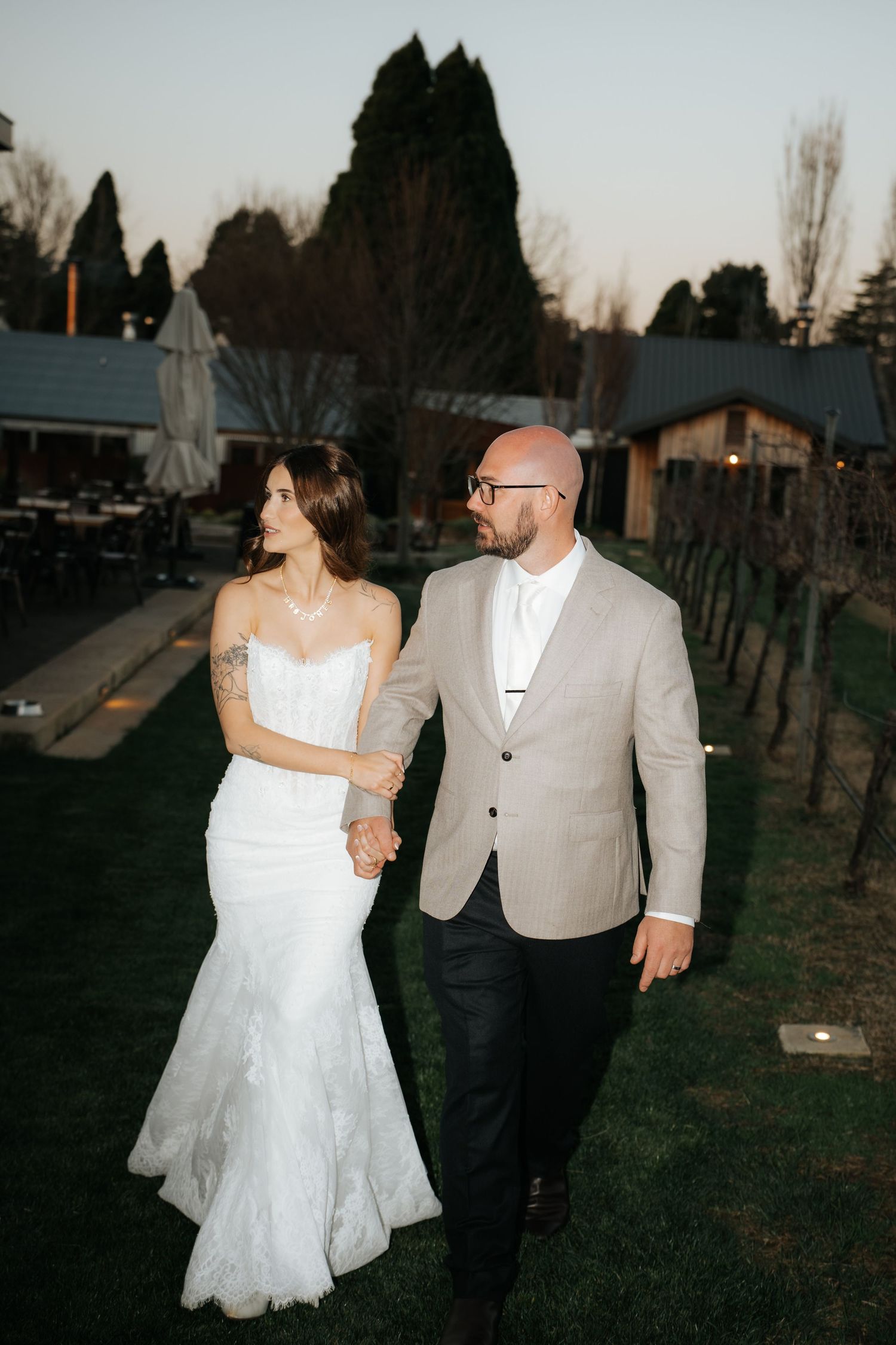 A bride in a white mermaid gown and groom in a tan blazer walk hand-in-hand at a vineyard wedding at dusk.