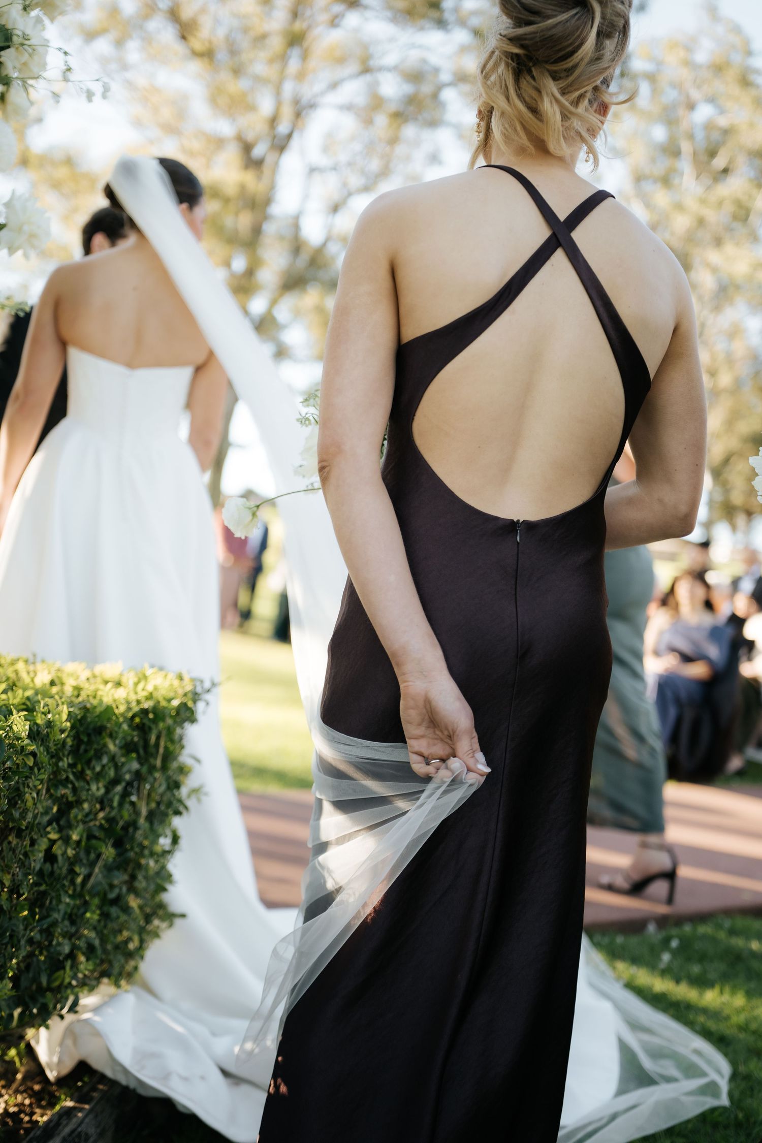 Bridesmaid in backless black gown holds bride's white veil train during outdoor wedding ceremony.