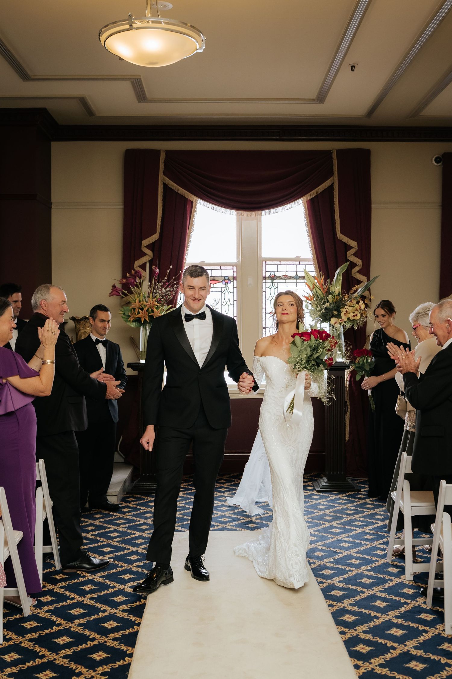 Bride and groom walk down the aisle hand in hand after wedding ceremony, guests applauding in elegant ballroom.