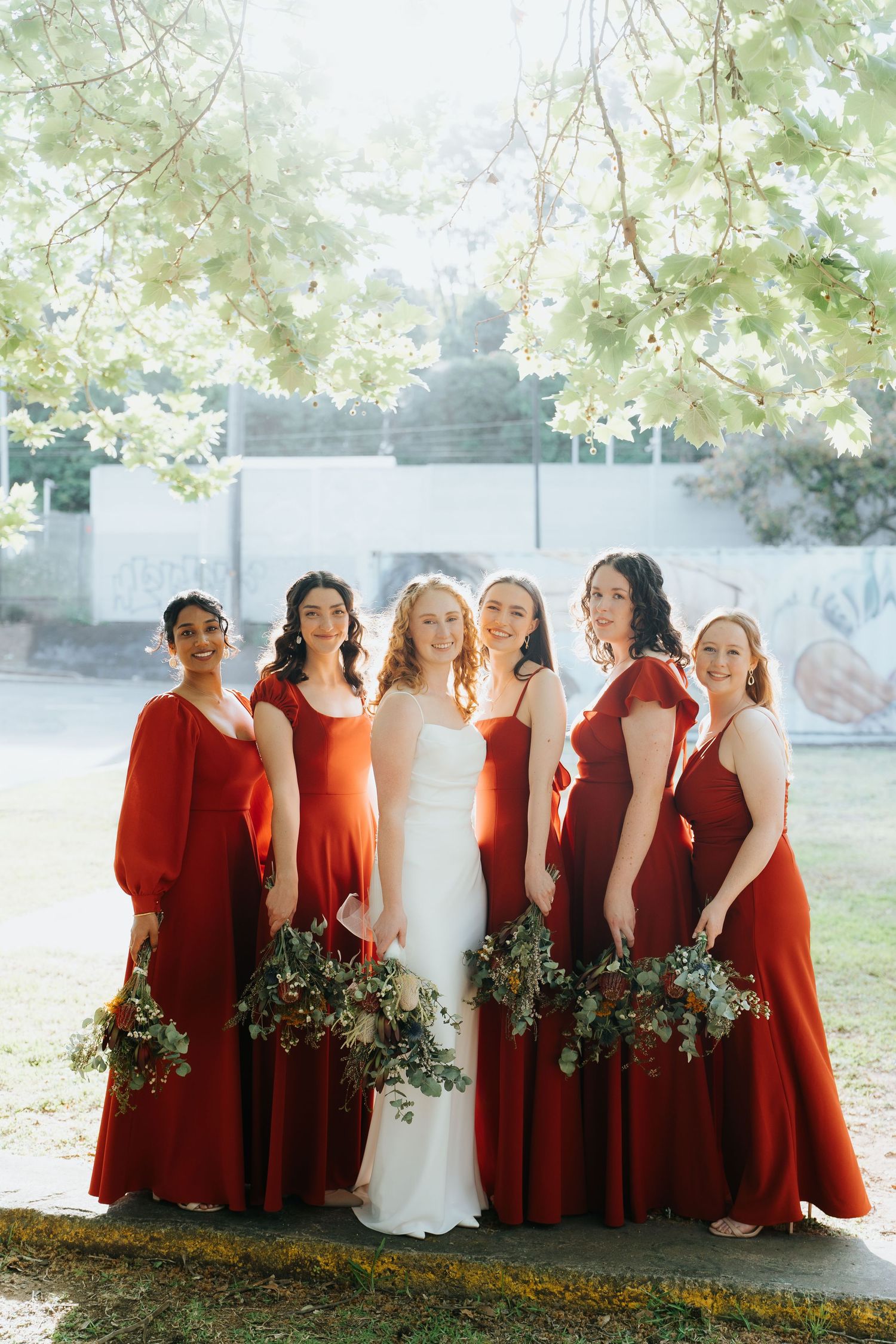 Bride in white gown poses with five bridesmaids in red dresses holding greenery bouquets outdoors.