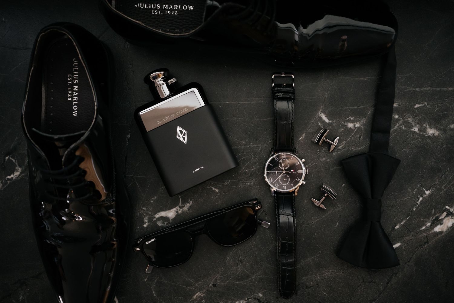 Dark moody flatlay of groom wedding accessories including watch, bow tie, cologne, cufflinks, and dress shoes.