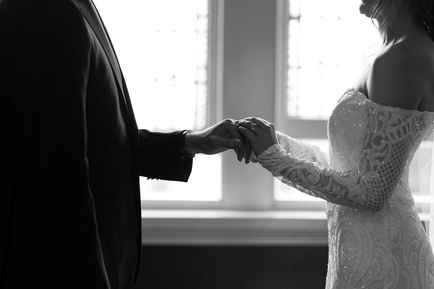 Black and white photo of bride and groom holding hands in wedding attire near a window.