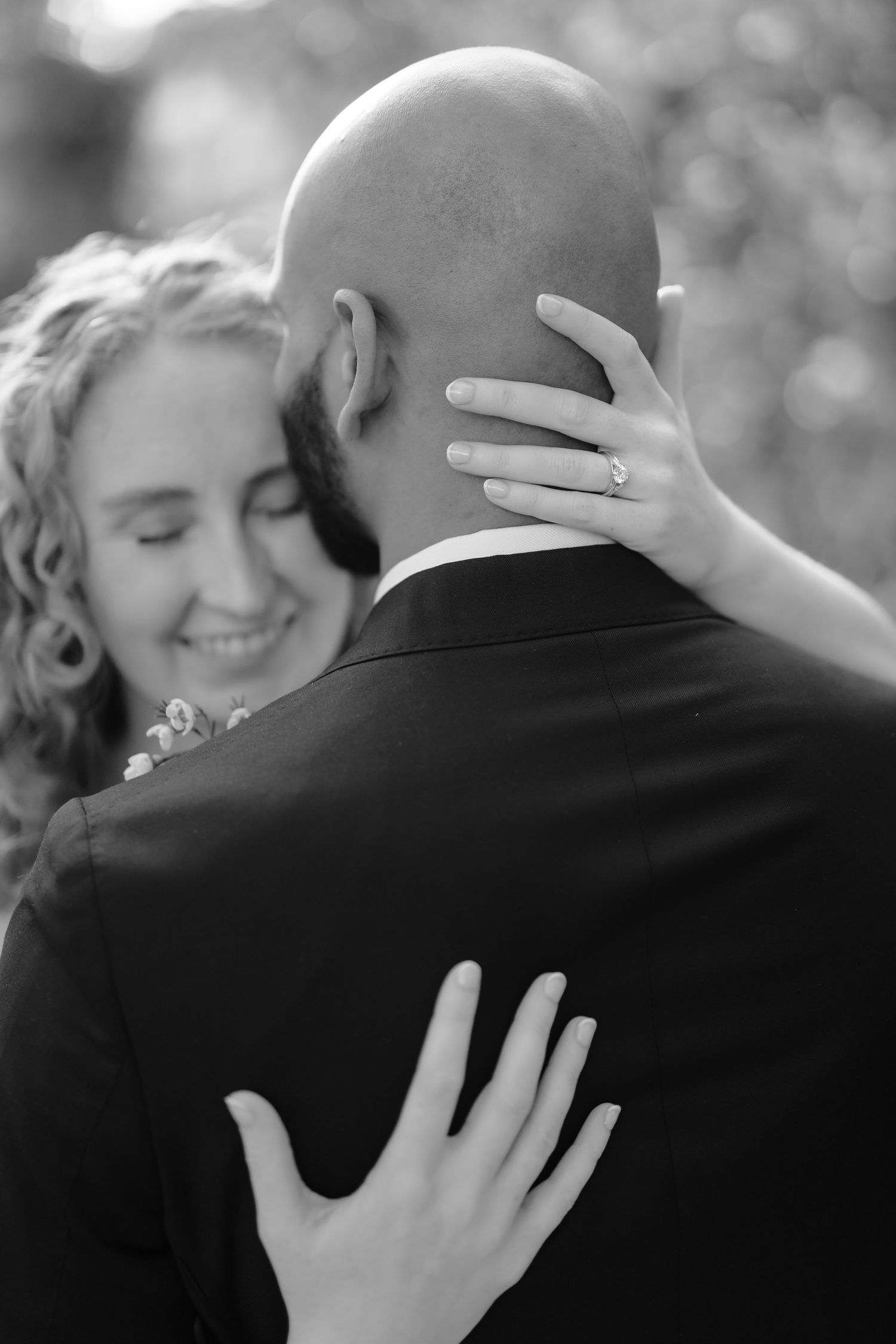 Black and white photo of smiling bride embracing bald groom, showcasing elegant diamond engagement ring on her hand.