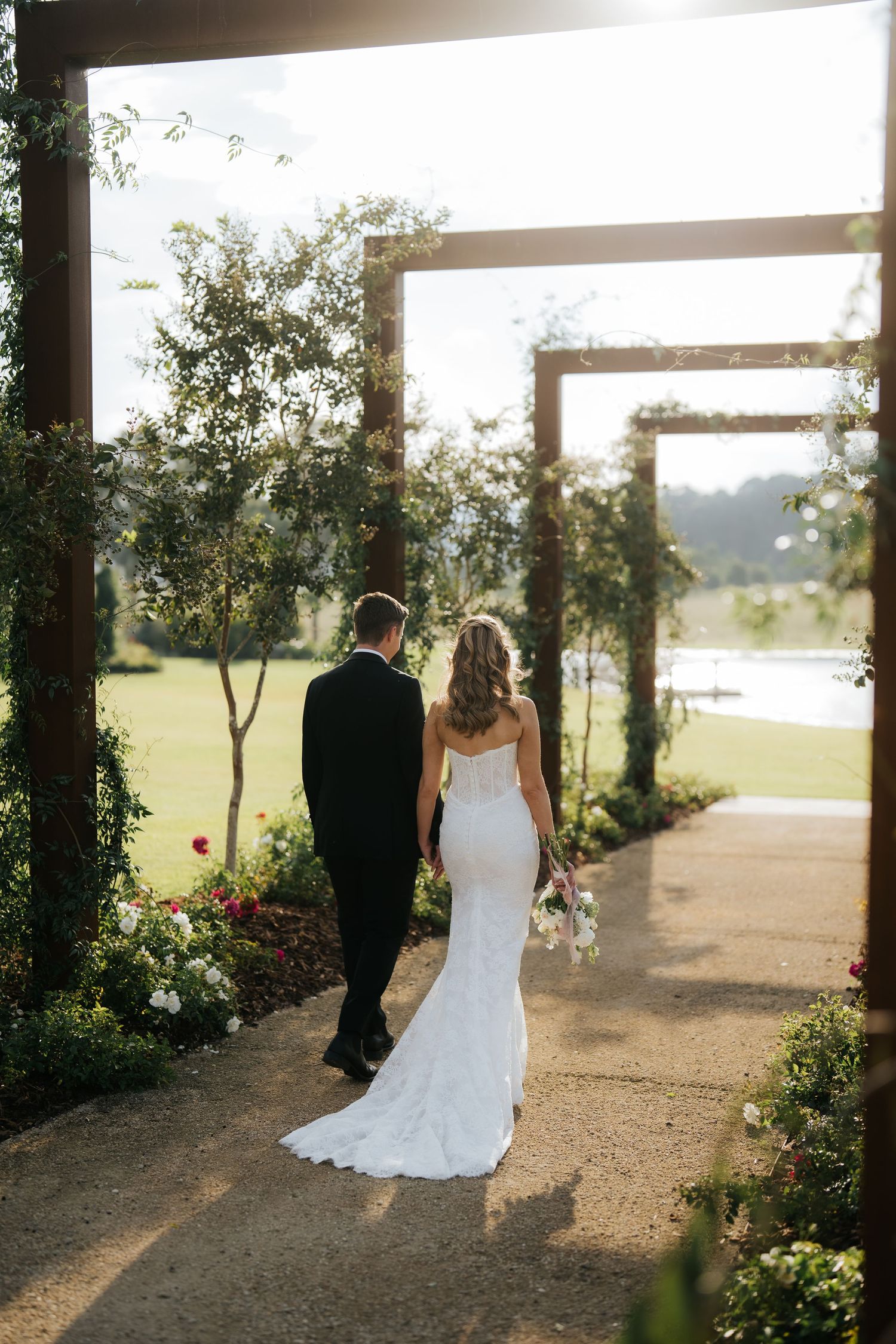 Bride and groom walking hand in hand through a floral garden pergola toward a sunlit lake on their wedding day.