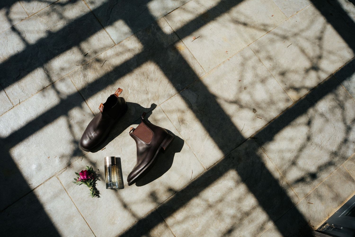 Brown leather Chelsea boots with small flowers and a bottle on marble tiles, with dramatic tree branch shadows.