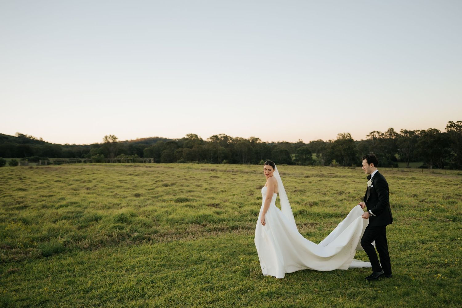 Groom in black tuxedo carrying bride's long white wedding veil and train across a green sunlit field.