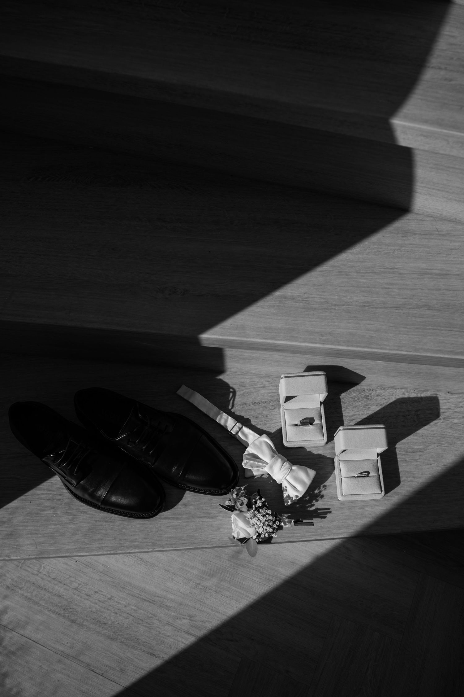 Black and white photo of groom wedding accessories including dress shoes, bow tie, boutonniere, and ring boxes on wooden steps.