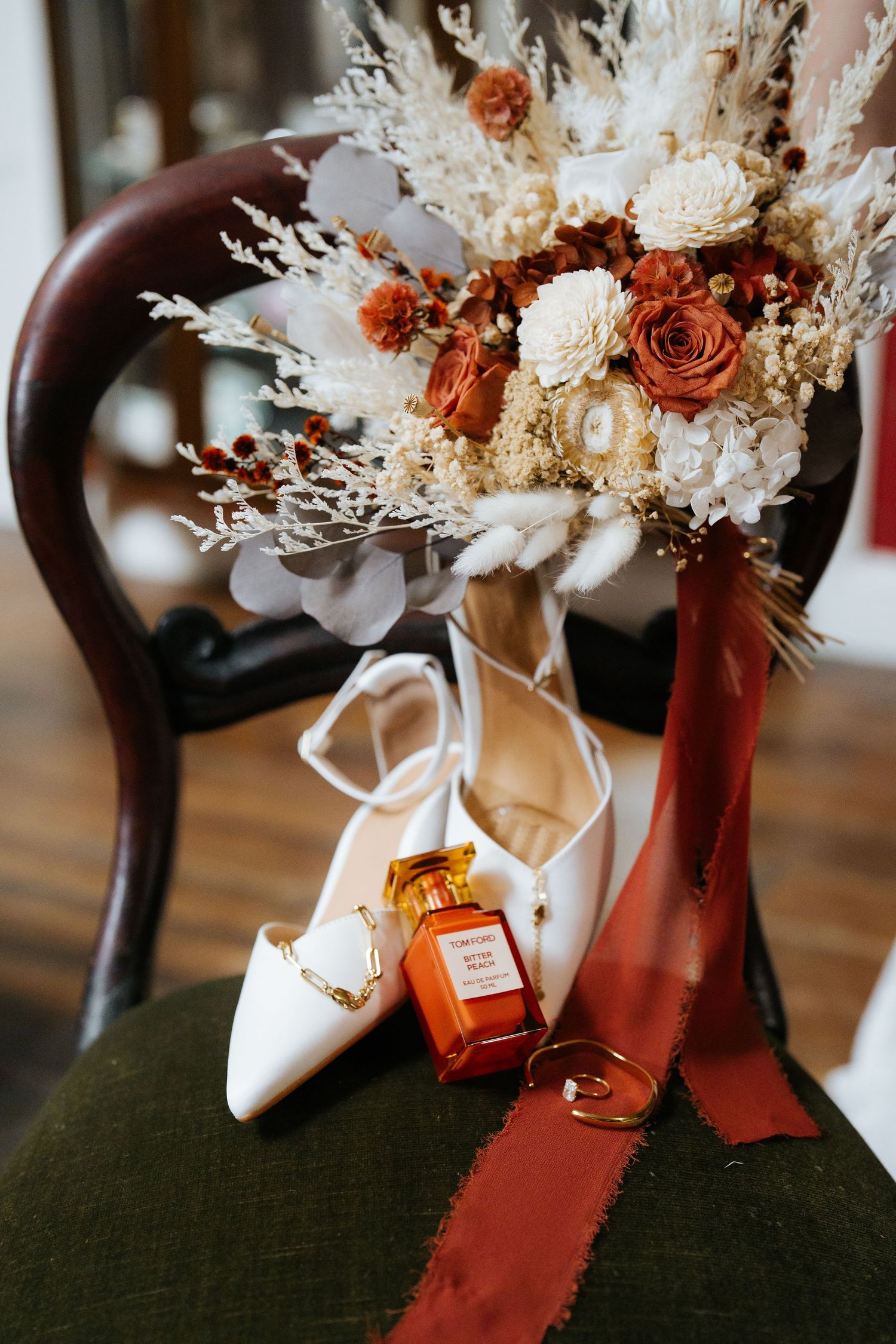 White bridal heels, gold rings, amber perfume bottle, and a dried floral bouquet with rust and cream tones on a chair.
