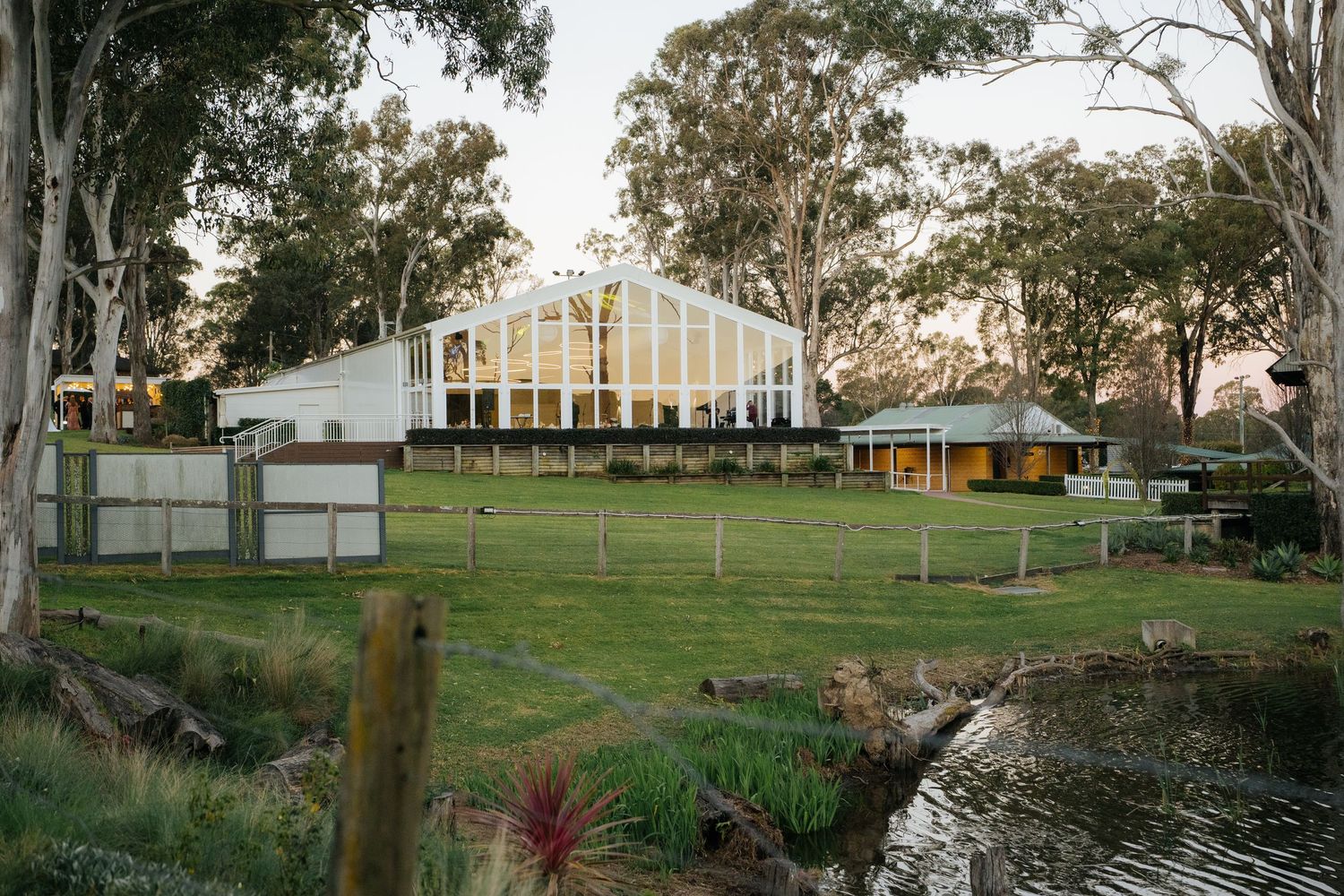 Rural Australian property with greenhouse, farmhouse buildings, lush green lawns, eucalyptus trees, and a small pond at dusk.