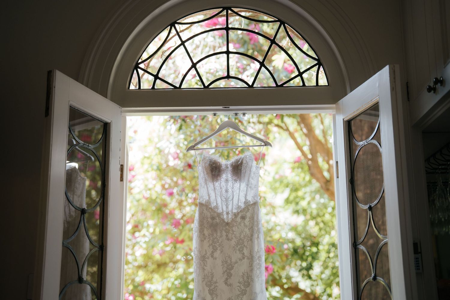 Elegant lace wedding dress hanging on double doors with stained glass transom window and garden view.