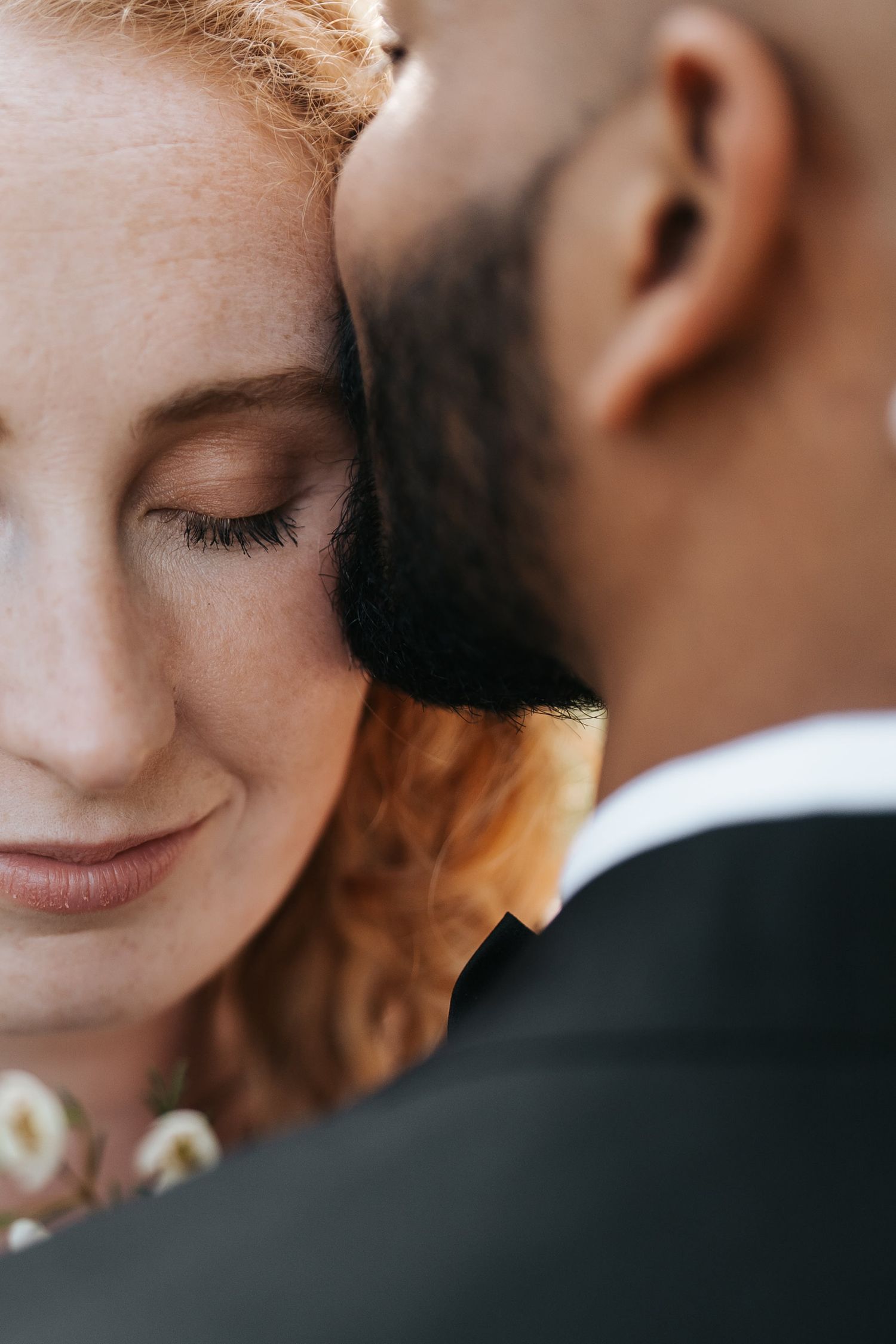 A tender moment between an interracial couple, redhead woman smiling with eyes closed, resting her head against a man in a black suit.