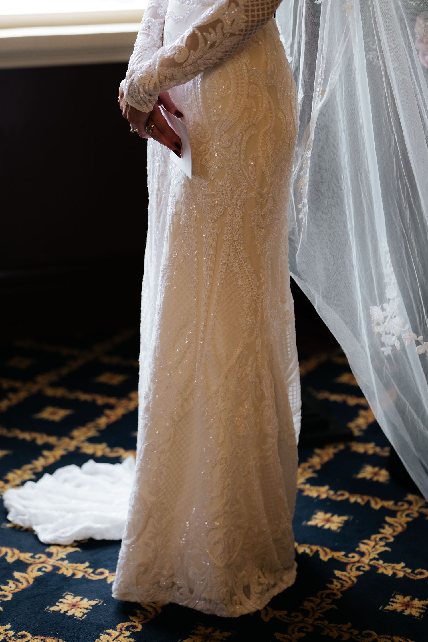 Bride wearing a fitted long-sleeve lace wedding gown with intricate beading and a flowing veil on patterned carpet.