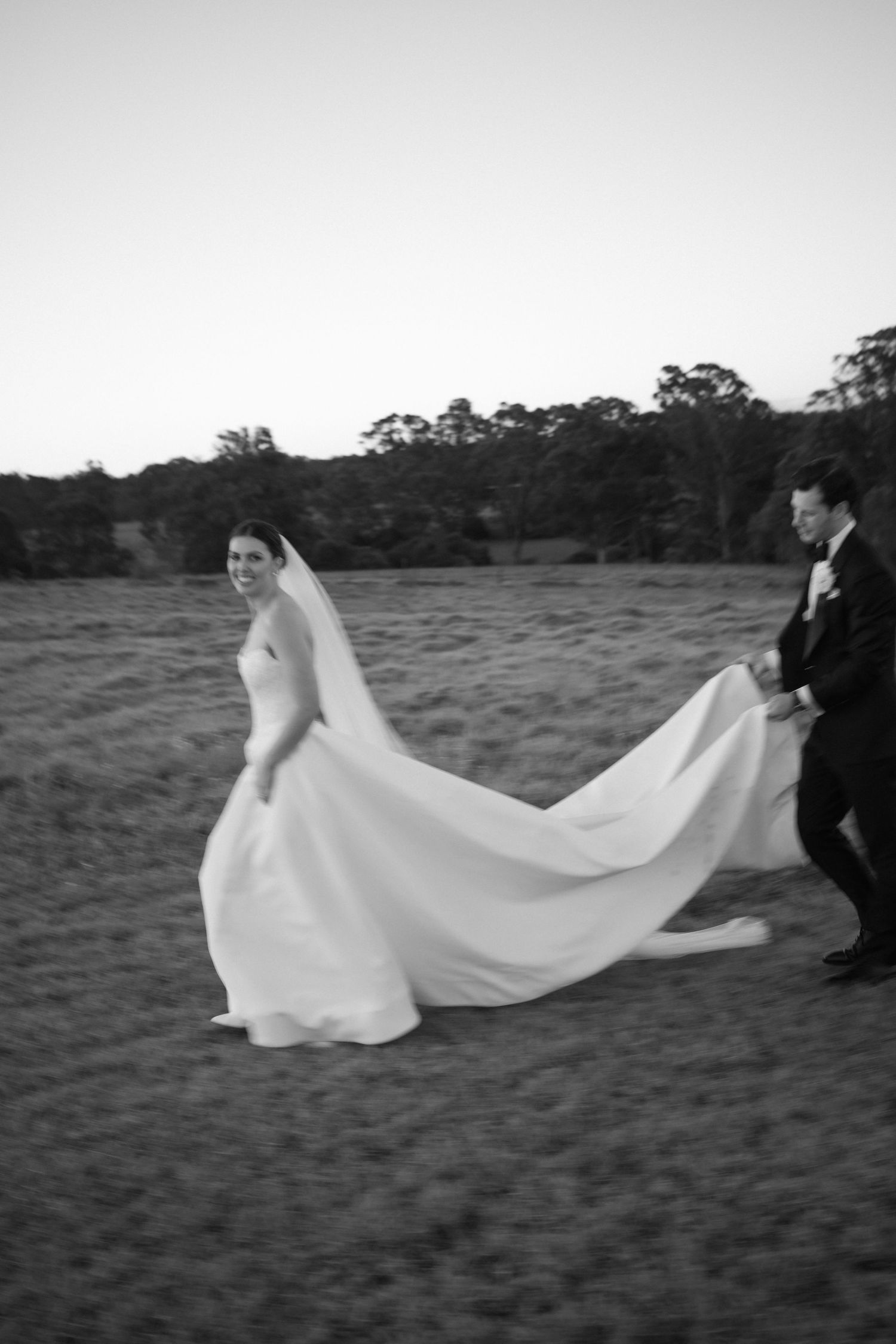 Black and white photo of bride and groom playfully running across field at dusk.