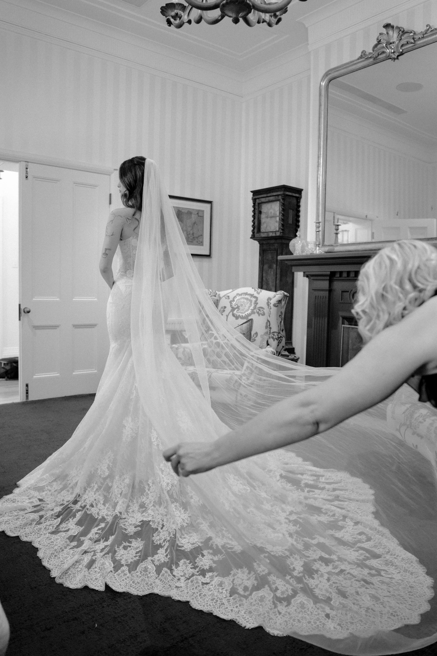 Black and white photo of bride in lace mermaid gown with cathedral veil being arranged by bridesmaid.