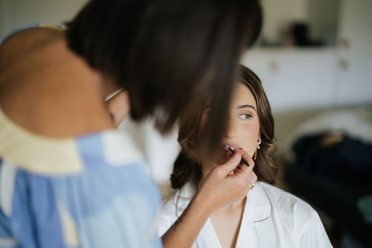 Makeup artist applying finishing touches to a bride in a white robe before her wedding ceremony.