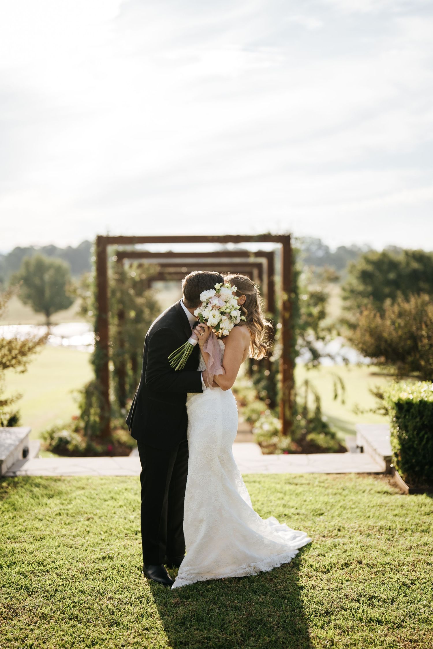 Bride and groom share a kiss outdoors at a garden wedding ceremony, with a rustic wooden arch and lush green lawn.