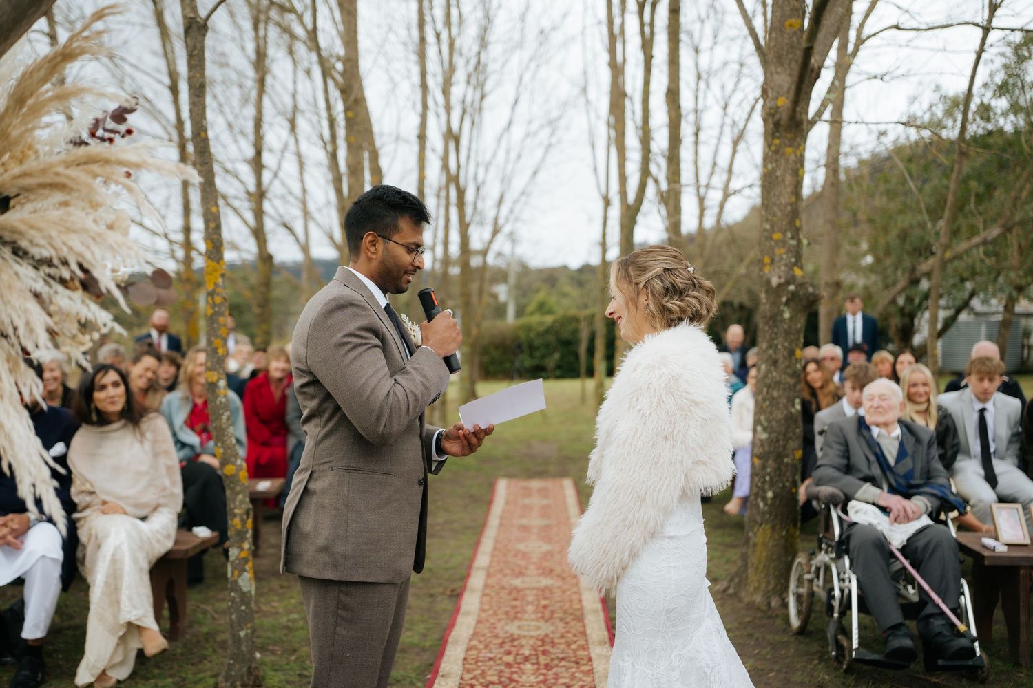 Groom reads vows to bride in white fur shawl during outdoor wedding ceremony with seated guests.