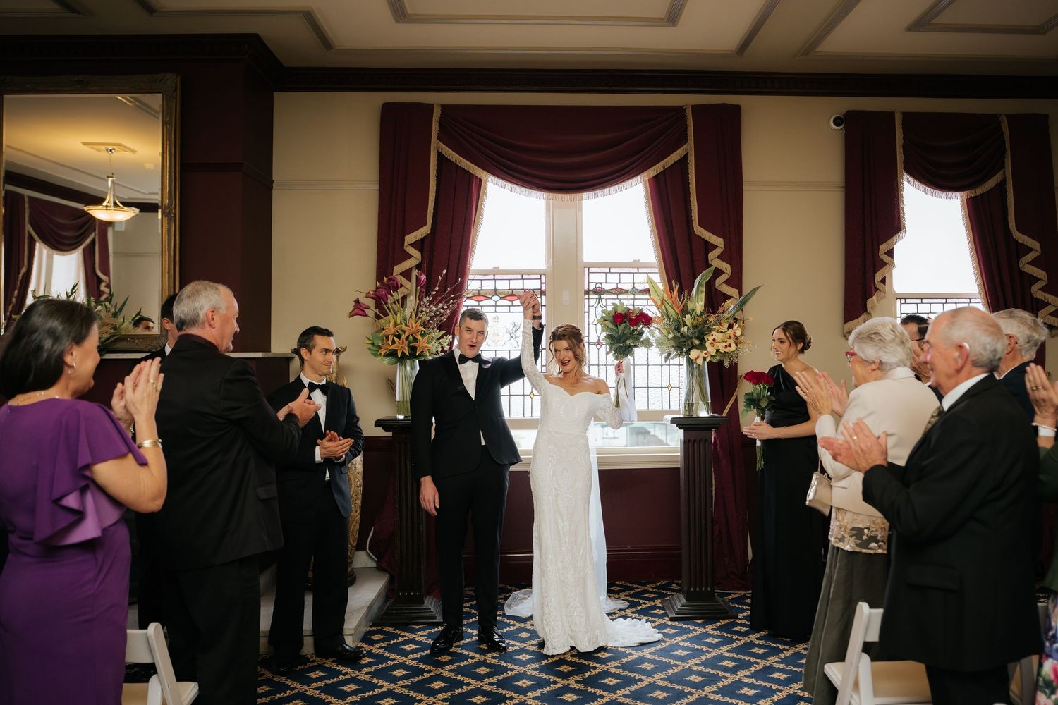 Bride and groom celebrate their recessional in an elegant ballroom, surrounded by applauding guests in formal attire.