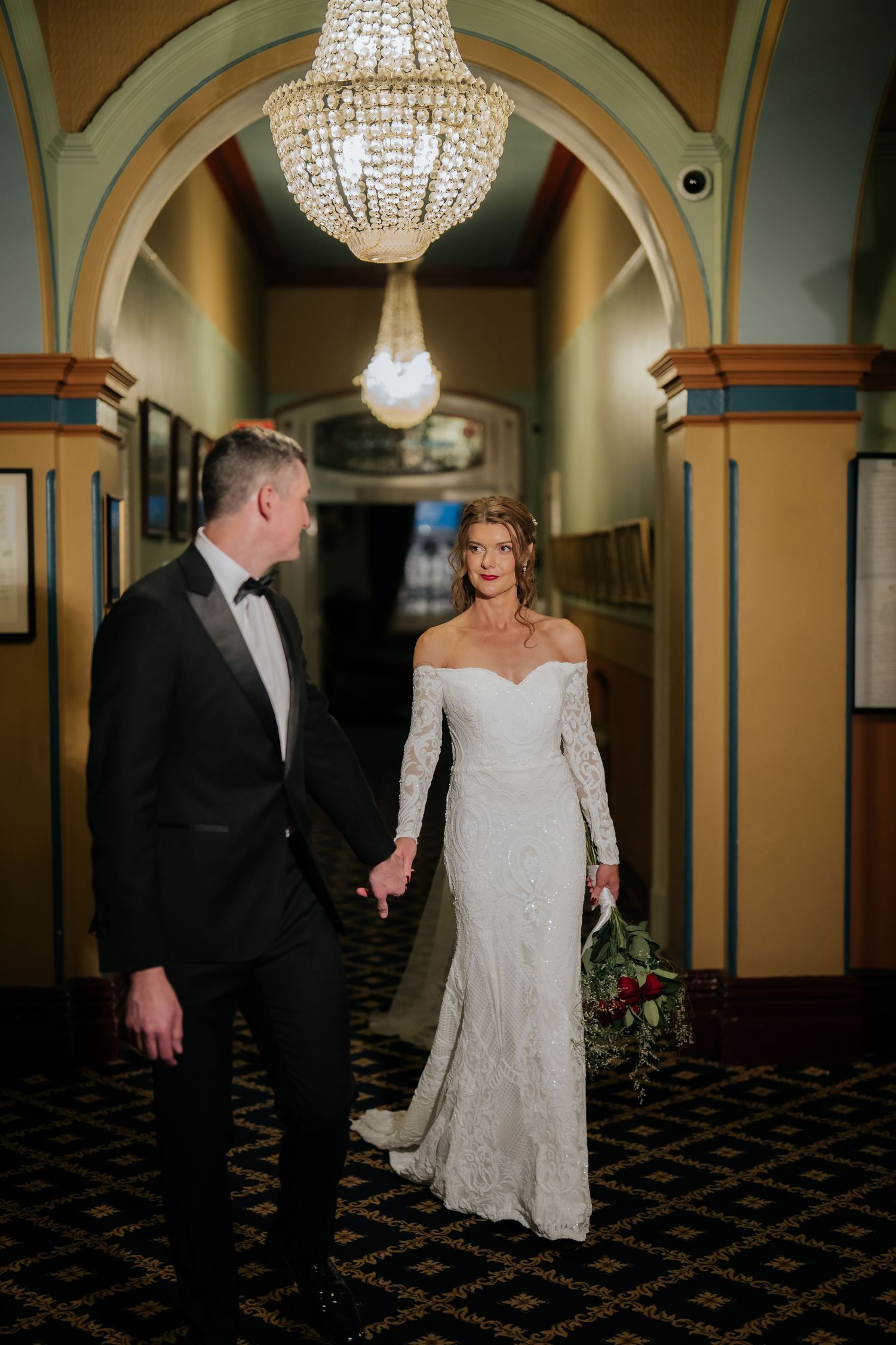 Bride in lace off-shoulder gown holds groom's hand in elegant chandelier-lit hallway.