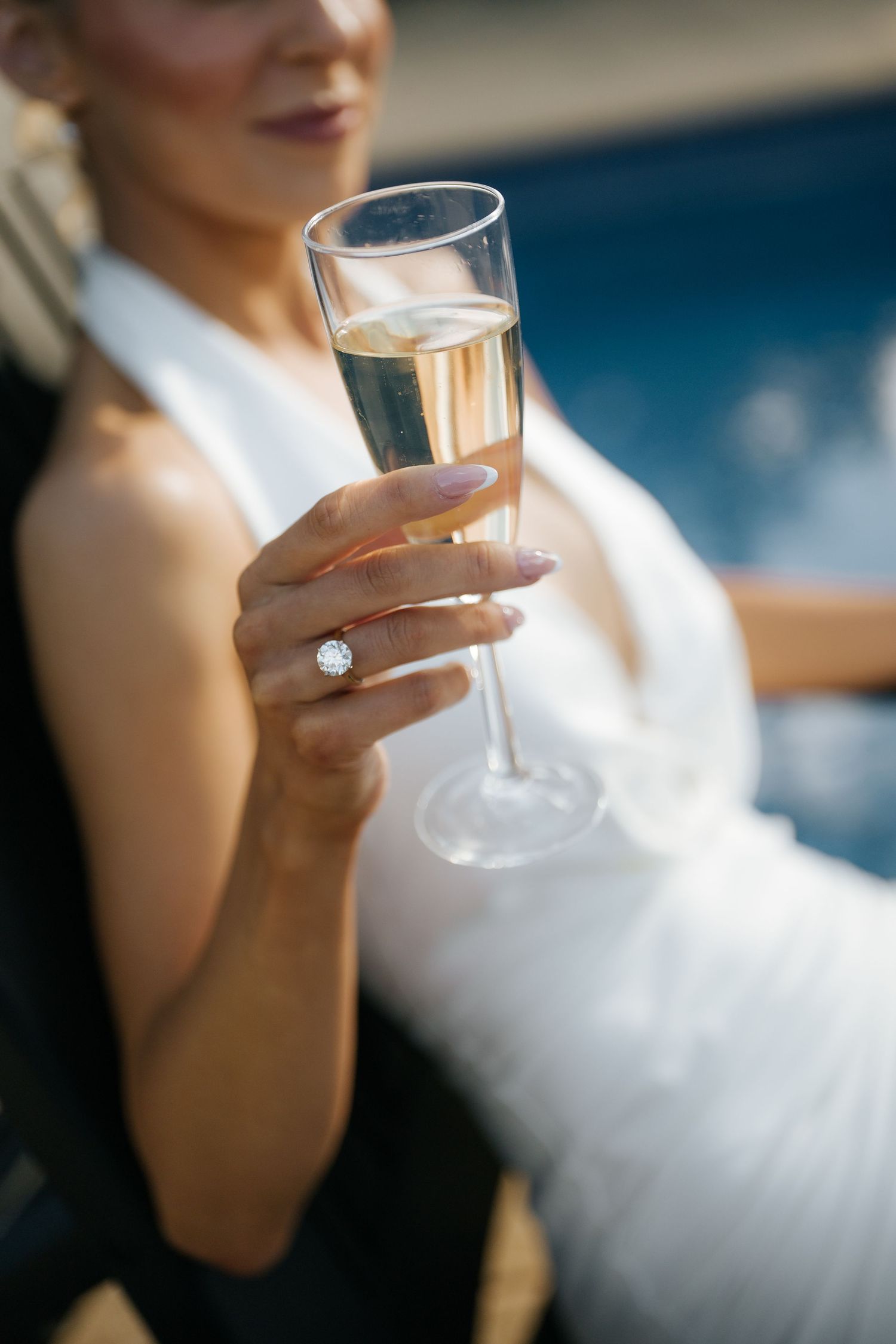Bride in white dress holding champagne flute with diamond engagement ring, celebrating near a pool.