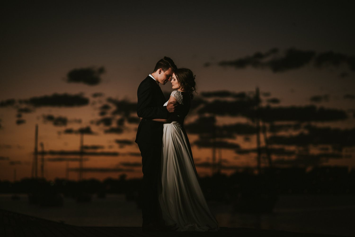 Bride and groom embracing at sunset with a dramatic orange sky and silhouetted boats in the background.