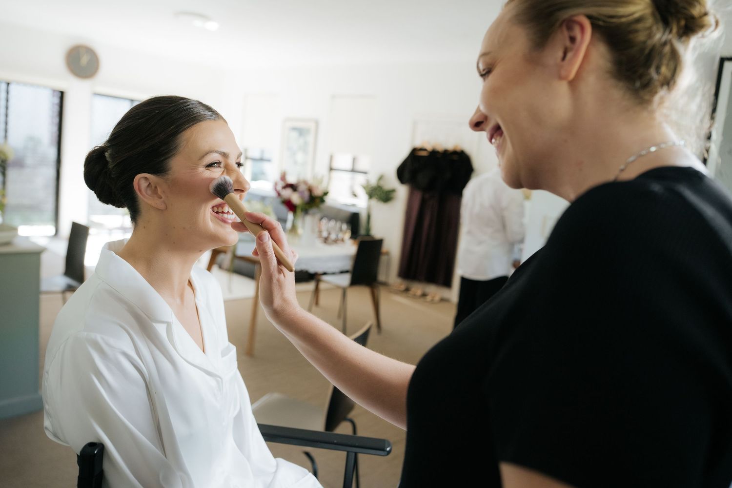 A makeup artist applying foundation to a smiling bride in a bright salon during wedding preparations.