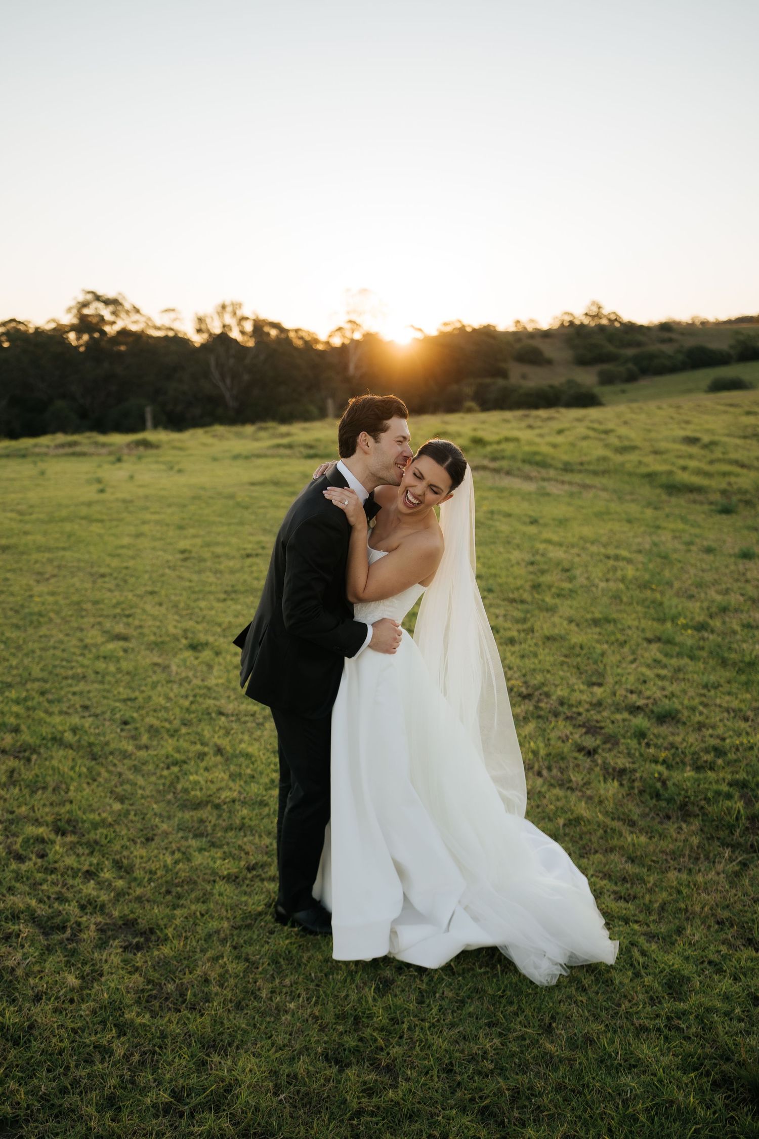 A bride and groom embrace at golden hour in a green field, the groom kissing her cheek as she laughs in her white gown.
